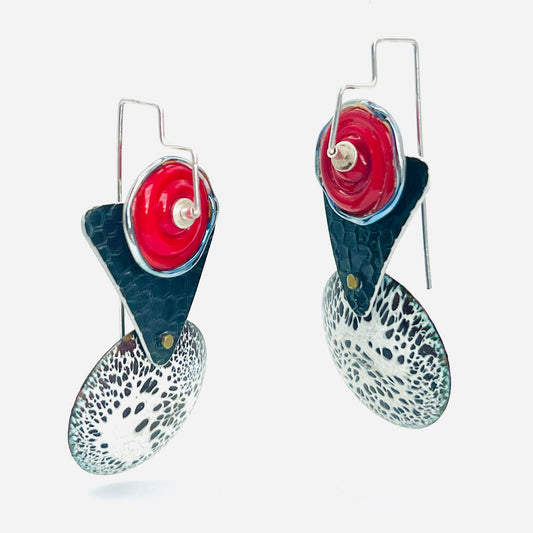 These earrings with their art deco vibe feature a bold balance of geometric shapes with an equally bold red, white and black color scheme that is sure to get noticed! A domed round disc shape in a black and white speckled pattern is done with the technique of vitreous enamel It is the foundation for these gorgeous earrings. A lamp work disc bead in bold red accents the top and has a lovely silver edge to it.